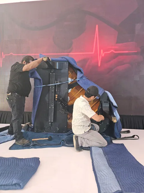 Movers working around the underside of a grand piano with protective blankets